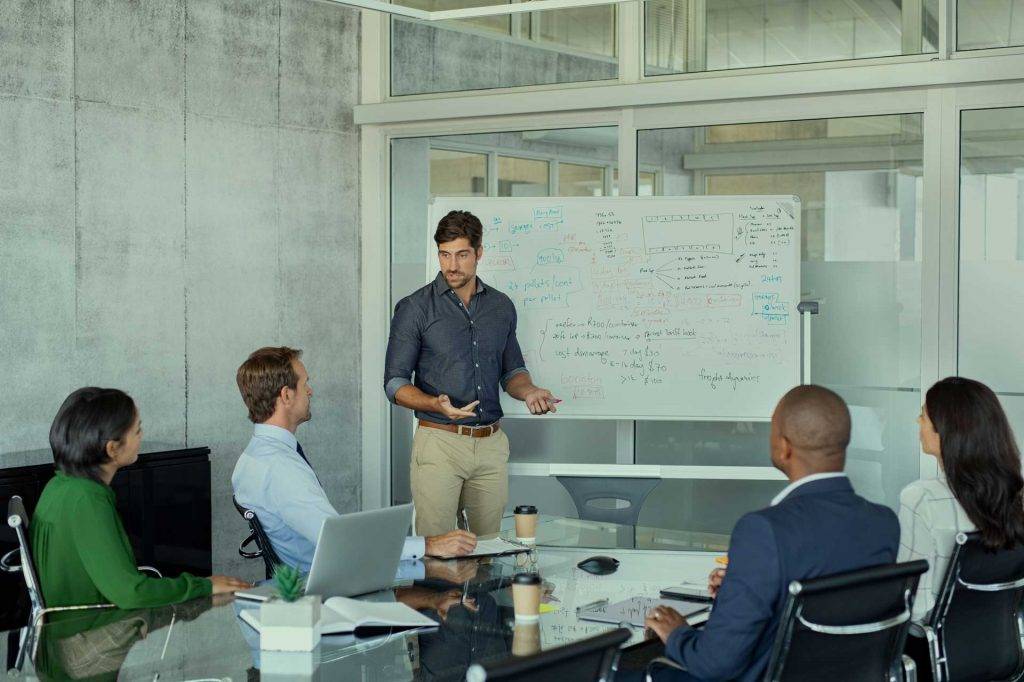 Teammeeting in einem modernen Büro: Ein Mann erklärt an einem Whiteboard, während vier Kolleginnen und Kollegen an einem Glastisch sitzen – Reinigung von Büroräumen und Industriereinigung von Officen