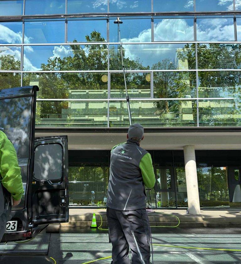 Reinigungsfachkraft säubert mit Teleskopstange die Glasfassade eines Gebäudes, im Vordergrund steht ein Transporter – Fassadenreinigung in Dresden