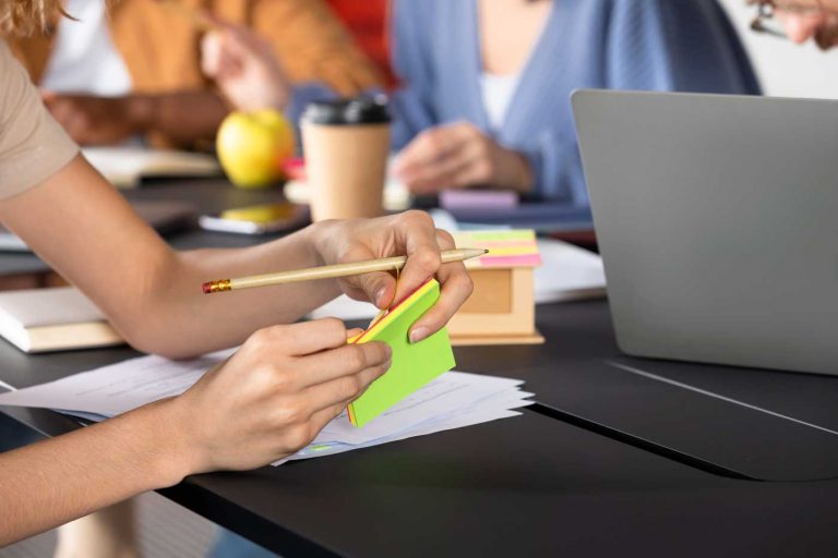 Hand schreibt auf grüne Haftnotizen am Schreibtisch mit Laptop und Team im Hintergrund – Reinigungsseminare und Schulungen für Immobilienverwalter