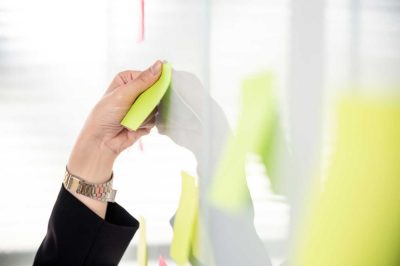 Klebezettel an Glaswand – Planung Hand klebt einen gelben Klebezettel an eine Glaswand – Planungsaufgaben in Reinigungsseminaren für Pflegeeinrichtungen