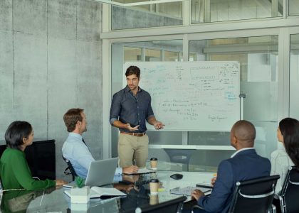 Teammeeting in einem modernen Büro: Ein Mann erklärt an einem Whiteboard, während vier Kolleginnen und Kollegen an einem Glastisch sitzen – Reinigung von Büroräumen und Industriereinigung von Officen