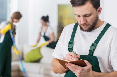 Teamführung Reinigung – Mann mit Klemmbrett Reinigungsmitarbeiter in grüner Latzhose notiert Informationen im Wohnzimmer, während Kollegin mit Wischmopp reinigt – Teamführung und DIN-Reinigungsseminare